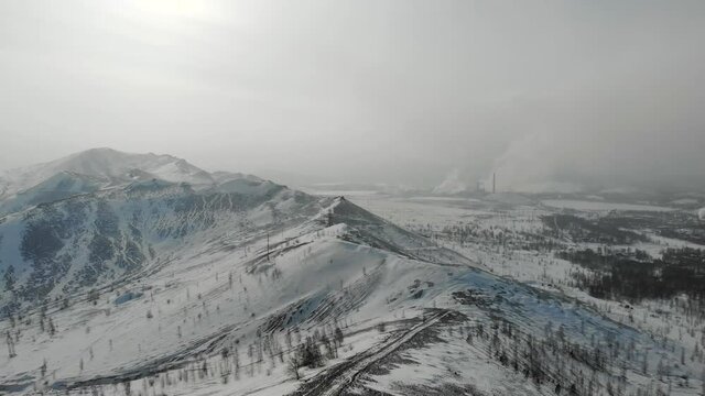 Aerial view of factory. The winter hills are covered with snow. Behind wires and electrical supports. Acrid poisonous smoke comes from the chimneys of the plant. Environmental pollution