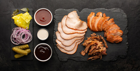 Assortment of smoked meats, pickled vegetables and various sauces on a slate board, top view. Meat...
