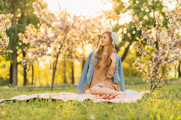 smiling young woman, with a bouquet of pink tulips in a straw hat, sits on the lawn in a blooming...