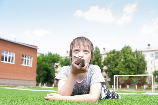 Young Boy Lies On Grass In Summer Park And Using Smartphone. Daily Life Child With Down Syndrome, Disability