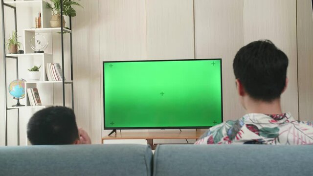 Father And His Son Are Watching Green Mock-Up Screen Tv While Sitting On A Couch At Home
