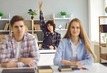 Smart teen schoolboy in casual wear sitting at classroom desk and raising hand in class. Active aspiring good high school student with curly dark brown hair asking or answering question in a lesson
