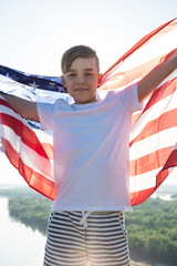 Blonde boy waving national USA flag outdoors over blue sky at the river bank