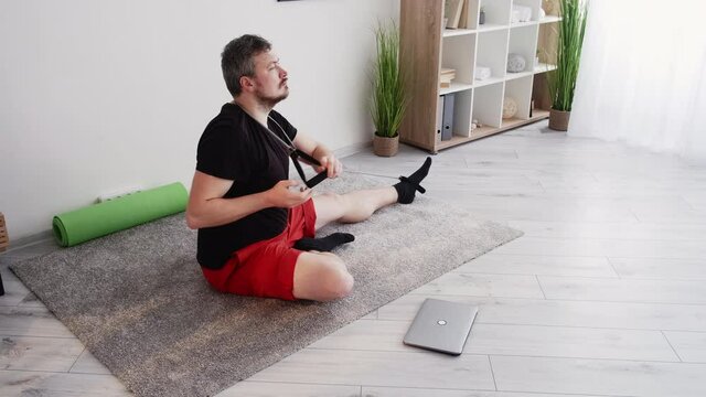 Home fitness. Performing man. Sport skill. Healthy lifestyle. Middle-aged funny guy sitting floor closing laptop doing resistance band workout different pose kissing biceps light room interior.