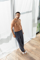 Obraz premium Shirtless man in pants and slippers looking at camera near window