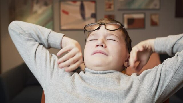 Portrait of small tired boy wearing circle glasses doing warm-up exercises, stretching his hands and back. Child rubbing his eyes after long study at computer during the coronavirus isolation at home.