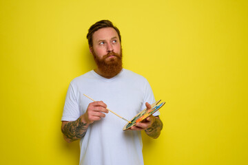 Undecided man with beard and tattoo is ready to draw with brushes
