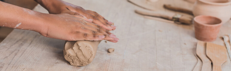 Fototapeta premium partial view of female african american hands rolling out piece of clay on wooden table in pottery, banner