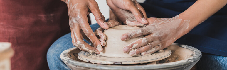 partial view of young african american couple modeling wet clay on wheel with hands in pottery,...
