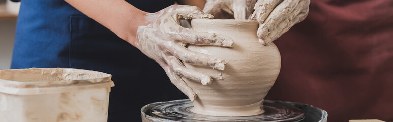 partial view of young african american couple shaping wet clay pot on wheel with hands in pottery, banner