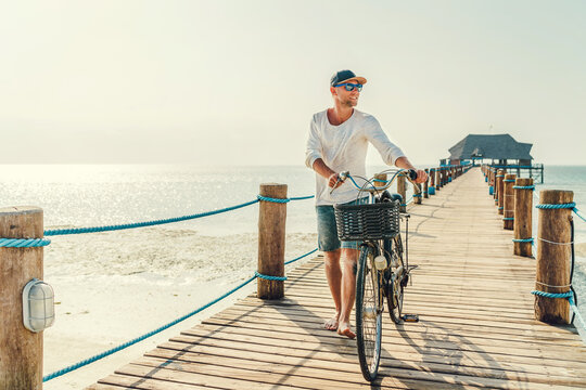 Portrait of a happy smiling barefoot man dressed in light summer clothes and sunglasses riding a bicycle on the wooden sea pier. Careless vacation in tropical countries concept image.