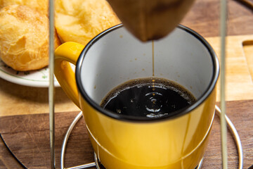 Café sendo coado no coador com uma caneca amarela abaixo com um prato de pão de queijo ao fundo.