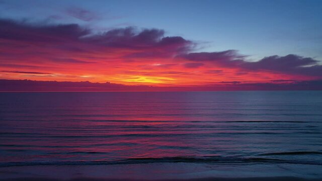 Aerial of dramatic predawn sky with colors of orange, red, yellow, and purple over a dark purple ocean.