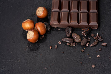 Close-up of a chocolate bar with nuts and cocoa beans on a dark stone background.