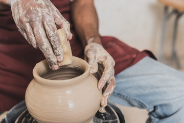 close up view of young african american man holding sponge and making wet clay pot on wheel in pottery