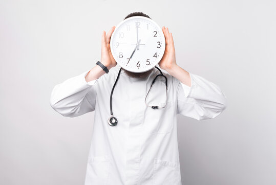 Photo Of Doctor With Clock In Front Of Face As Sign Of Time Pressure. Isolated On White Background