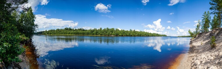 Obraz premium View from the sandy shore to the wide calm river on a clear summer day. Larger panorama of wide calm river on sunny summer day. White clouds are reflected in the blue water. 