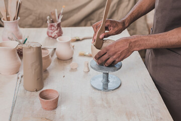 partial view of young african american man sculpting clay pot with spatula near table with equipment in pottery