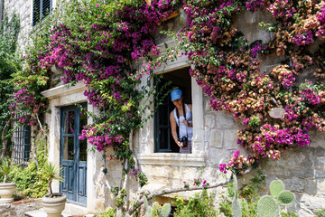 Fototapeta na wymiar Young girl looking through the window in old house full of flowers. Bougainvillea glabra in the wall of old house