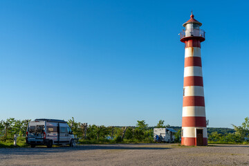 gray camper van parked next to a red and white lighthouse on the Danish coast undr a cloudless blue sky
