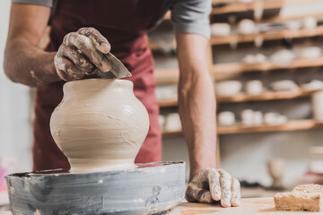 partial view of young african american man in apron shaping wet clay pot on wheel with scraper in pottery