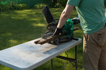 Man cutting up boards for disposal after home DIY project; miter saw set up on folding table in backyard