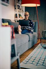 An attractive old woman with short hair sitting on the couch at her home and using laptop.
