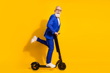 Full body profile photo of funny grey beard aged man ride scooter wear spectacles blue suit isolated on yellow background