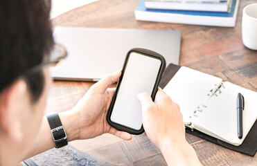 Man using smartphone with blurred background of laptop and books at wooden table work space, depth of field. Concept of remote work or study, work from home via online technology. Mockup mobile screen