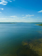 View from the drone is of a blue lake with a reflection of the sun and a green forest in the distance. Beautiful landscape from a drone on a sunny day. Vrtical photo