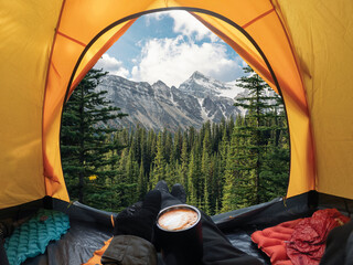 Traveler lying and holding coffee cup in yellow tent and enjoy the view of forest and mountain in...
