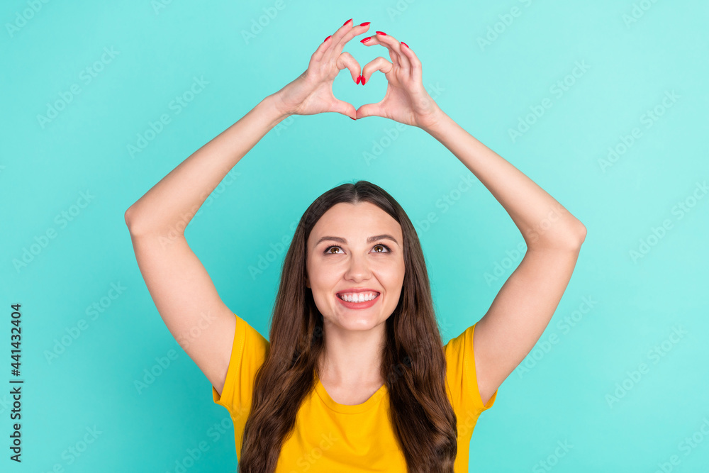 Sticker Photo of optimistic young lady show heart by hands look up wear yellow t-shirt isolated on vivid teal color background