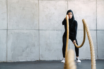 Young muslim woman in hijab working out with battle ropes indoors