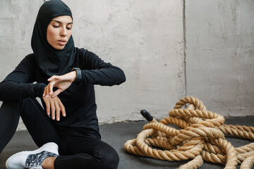 Young muslim woman in hijab looking at smartwatch while sitting on floor