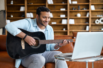 Obraz premium Smiling indian young man learning how to play guitar online, sitting on the sofa in front of the laptop, watching video tutorial, repeats exercises. E-studying and hobby online