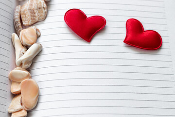 Two woven bright red hearts lying on the page of an open diary or glider in a line with seashells...