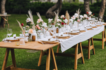 A large wooden, long, decorated with flowers, reeds, empty dishes, plates, forks, knives, glasses with a white tablecloth stands on the green grass in the forest. Wedding banquet in nature.
