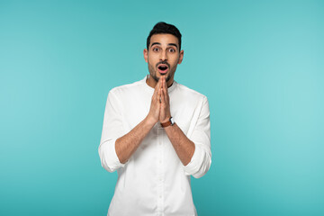 Amazed arabian man with praying hands looking at camera isolated on blue