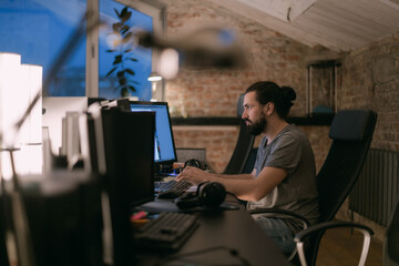 A young man works in the office in front of monitors in the evening.