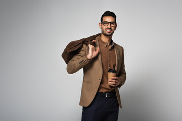Fototapeta premium Smiling arabian businessman holding coffee to go and handbag isolated on grey