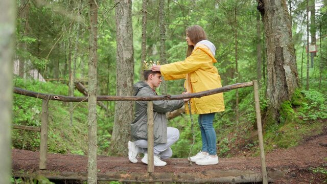 Tilt up side view of childish young couple of hikers having fun on wooden bridge over creek in woodland. Woman coronating and knighting her kneeling boyfriend. Guy with stick running after girlfriend