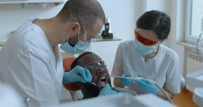 The African American male patient having teeth cleaning by the male dentist who is using a dental brush and mirror while a female assistant is using a saliva ejector and dental mirror.