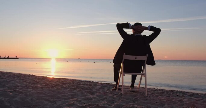 Man silhouette in black suit stretches arms sitting on wooden chair on sandy beach against orange sunset over endless sea