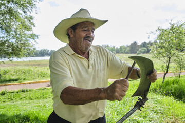 Hispanic male farmer sharpening an old scythe in cutting weeds on a farm