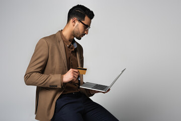Side view of arabian businessman holding credit card and laptop isolated on grey
