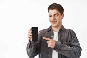 Close up of cheerful caucasian guy pointing finger at smartphone, showing mobile phone screen app, interface application, standing against white background