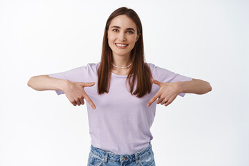 Portrait of cute happy woman pointing down, smiling white teeth and showing advertisement below, point at direction logo or banner, standing isolated against white background