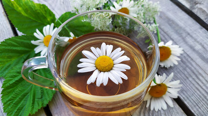 Obraz na płótnie Canvas Chamomile tea in a glass cup. Floral background. A bouquet of wildflowers and raspberry leaves in the background. A transparent cup with a chamomile drink stands on a wooden background. 