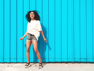 Obraz premium Beautiful black woman with afro curls hairstyle.Smiling hipster model in white t-shirt. Sexy carefree female posing in the street near blue wall in sunglasses. Cheerful and happy