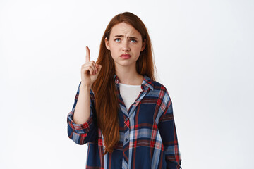Skeptical redhead girl points at something lame, grimacing and frowning upset, pointing and looking...